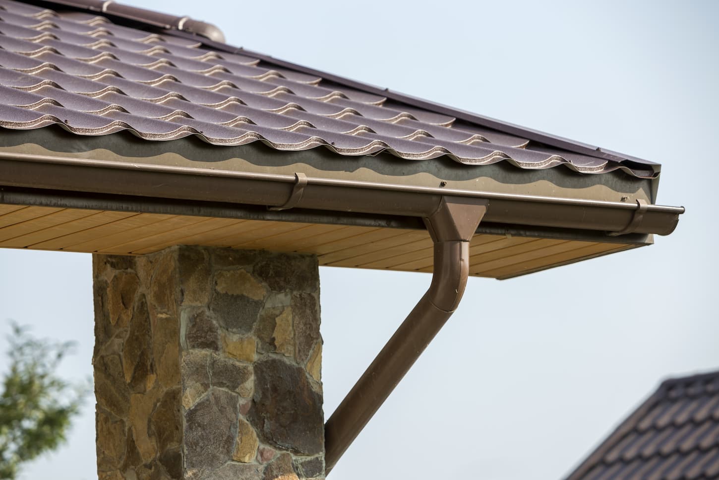 An image of a new modern house cottage corner with stucco walls decorated with natural stones, a brown shingled roof, and a rain gutter pipe system on blue sky copy space background.