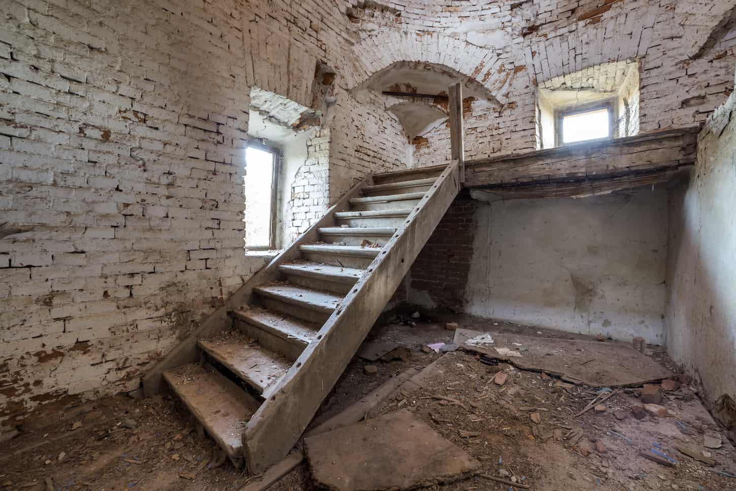 An image of a Large spacious forsaken empty basement room of an ancient building or palace with cracked plastered brick walls, small windows, a dirty floor, and a wooden staircase ladder.