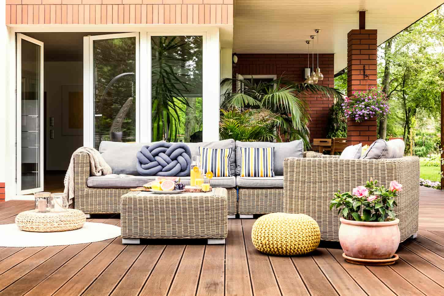 An image of Beige garden furniture with striped pillows on a wooden terrace with pink flowers and poufs.