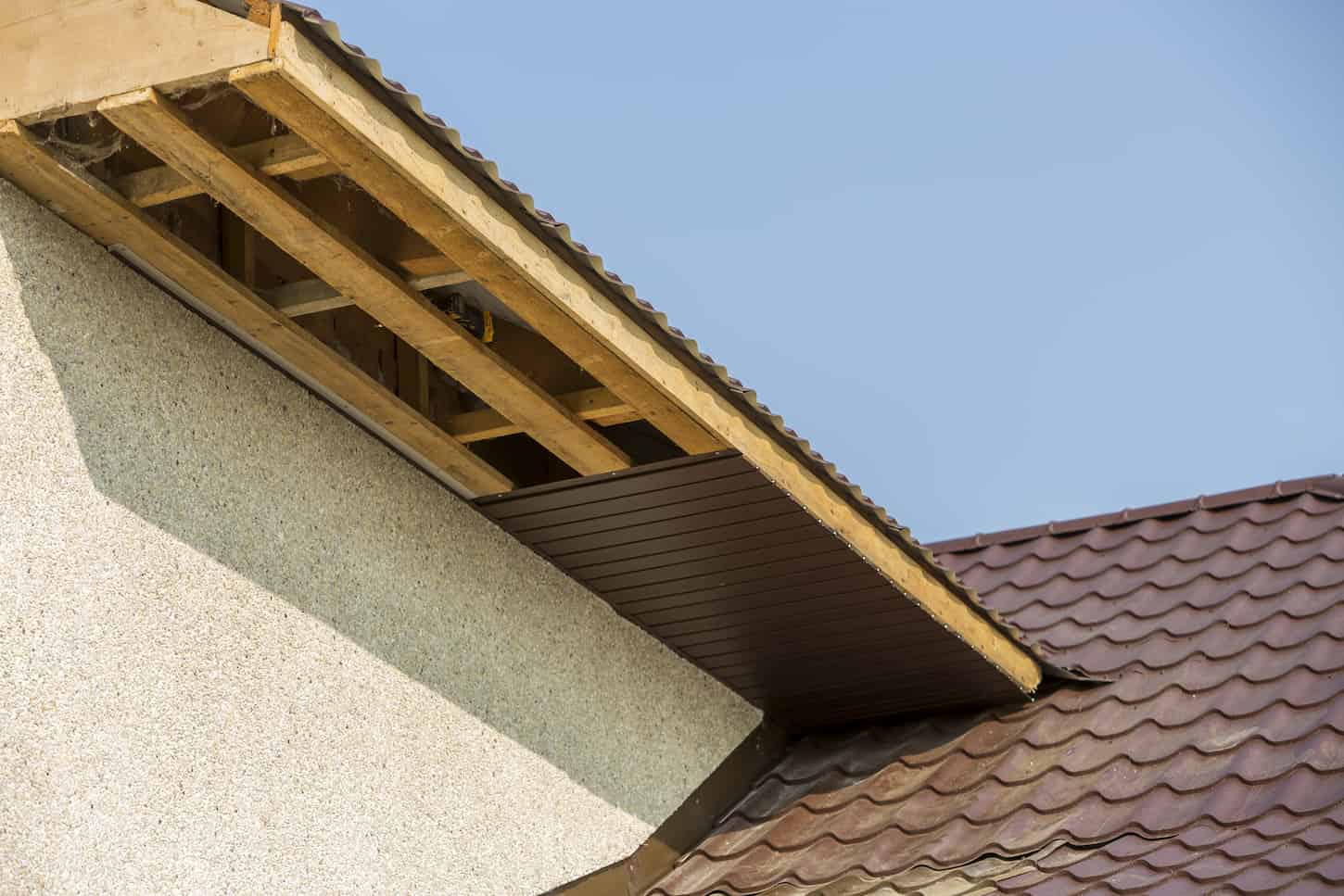 An image of the Bottom view detail of a new modern house cottage corner with stucco walls, brown shingled roof, and unfinished siding installation on clear blue sky copy space background. Real estate concept.