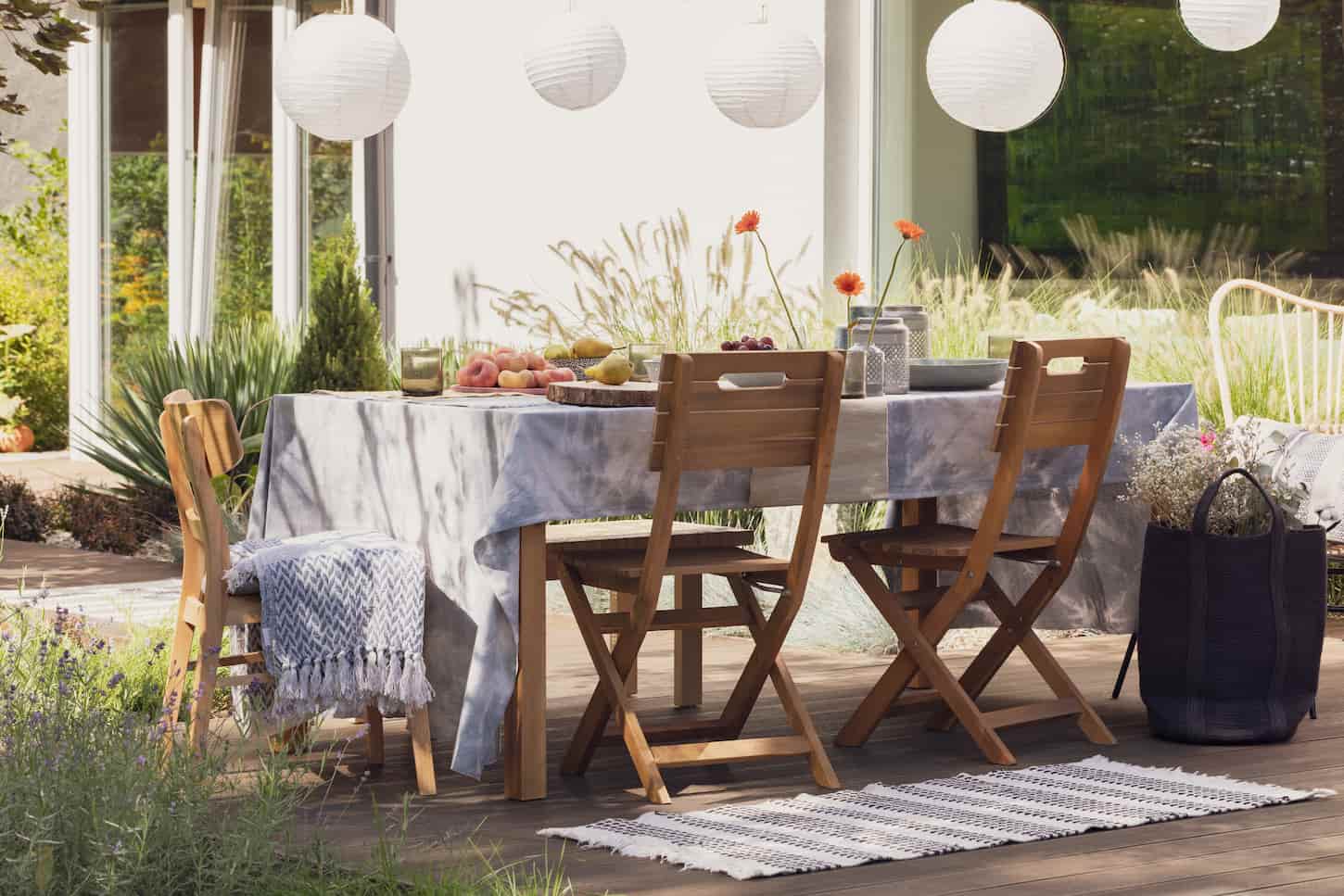 An image of a Rug next to wooden chairs at a table with food on the terrace with lamps and flowers.
