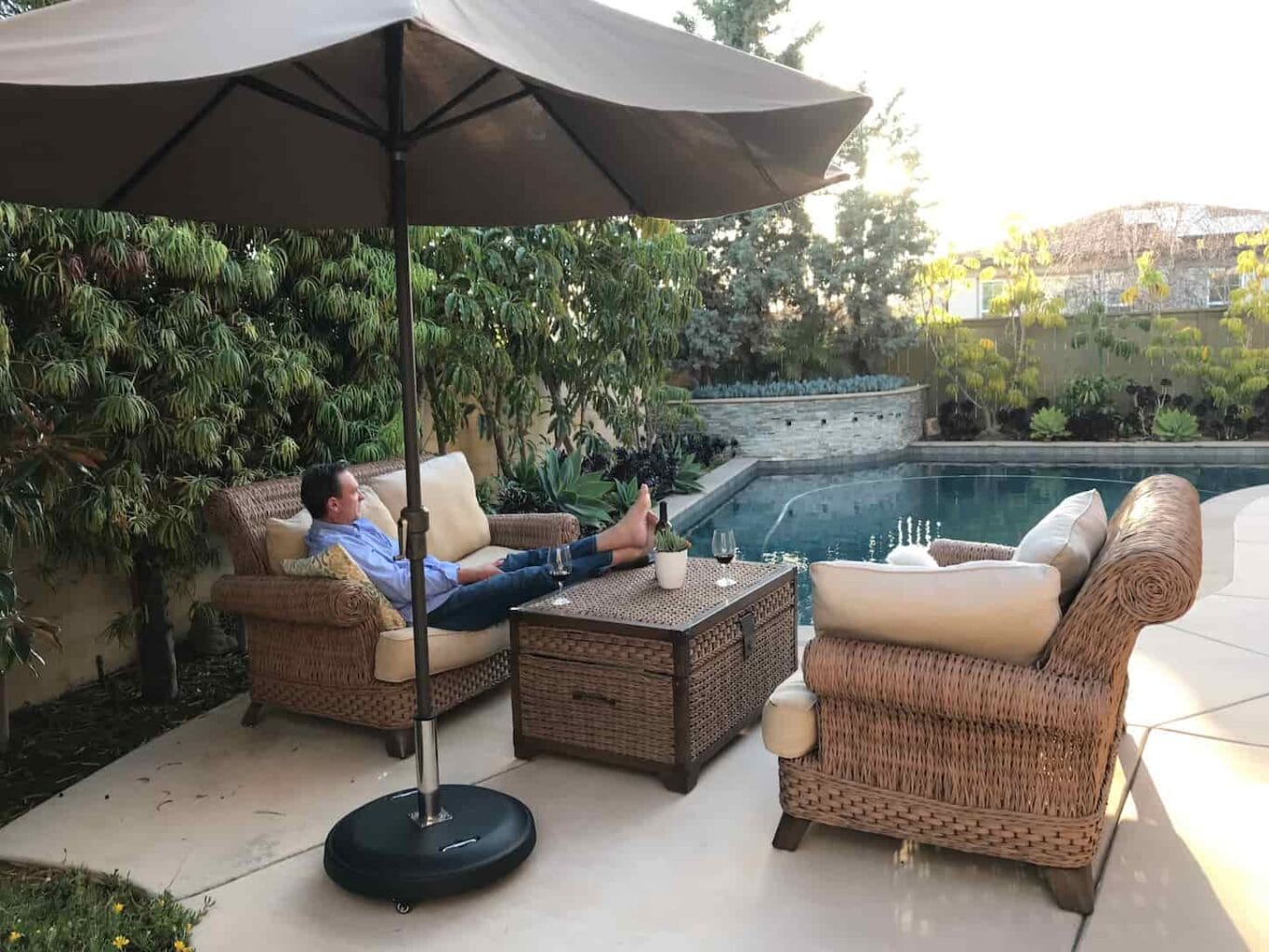 An image of a man sitting on outdoor patio furniture by the pool relaxing under an umbrella.