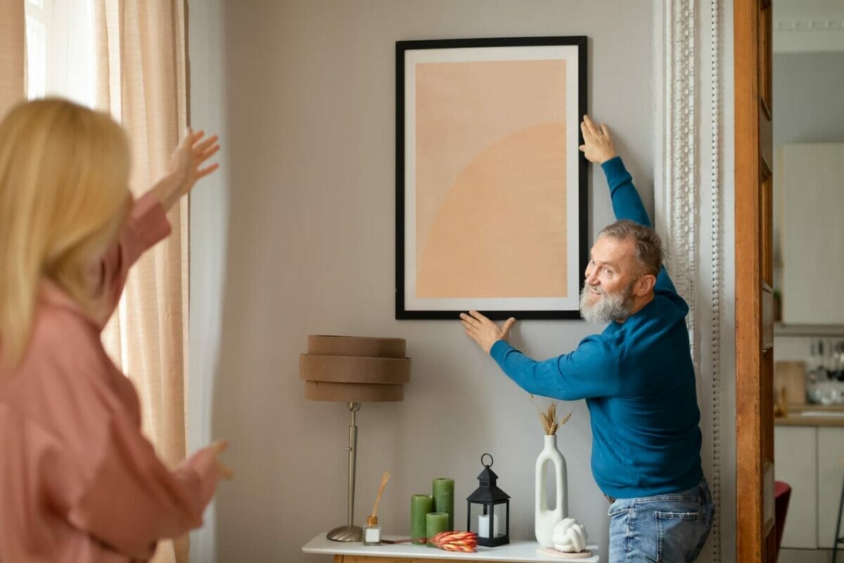 An image of Senior Spouses Hanging Poster In Frame On Wall Decorating Home.